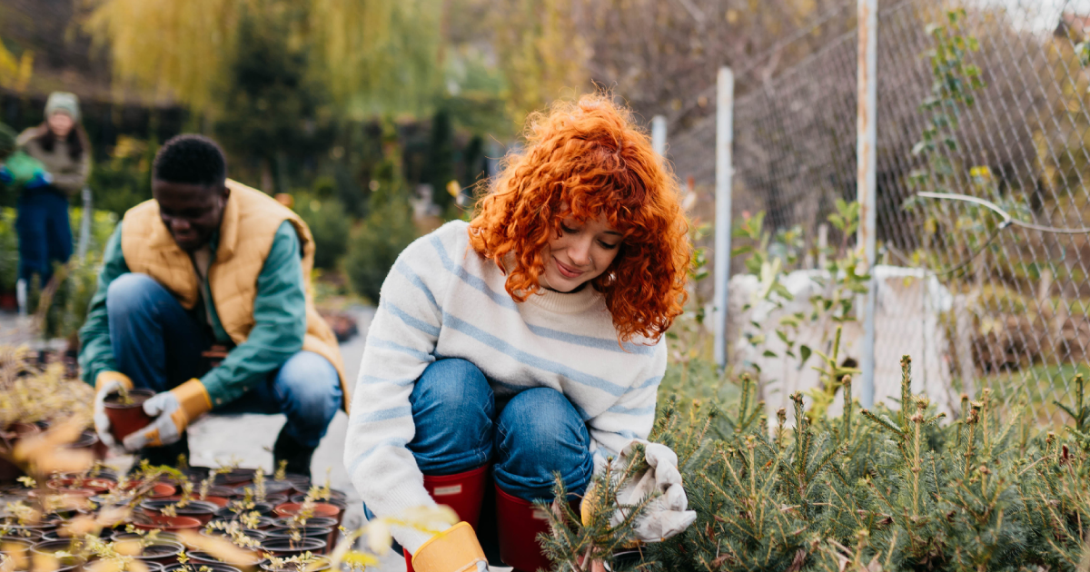 A young woman is smiling while gardening in a community garden, with two other youths helping out of focus behind her