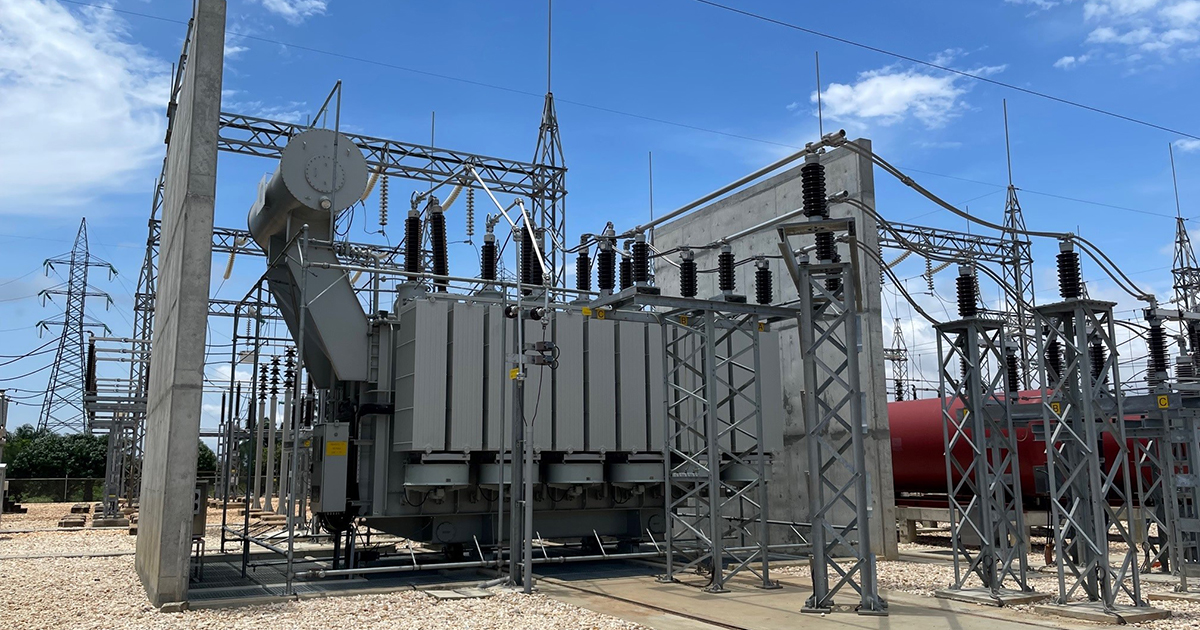 Large electrical power transformer and associated high-voltage equipment at an outdoor electrical substation, with transmission structures and power lines visible under a blue sky.