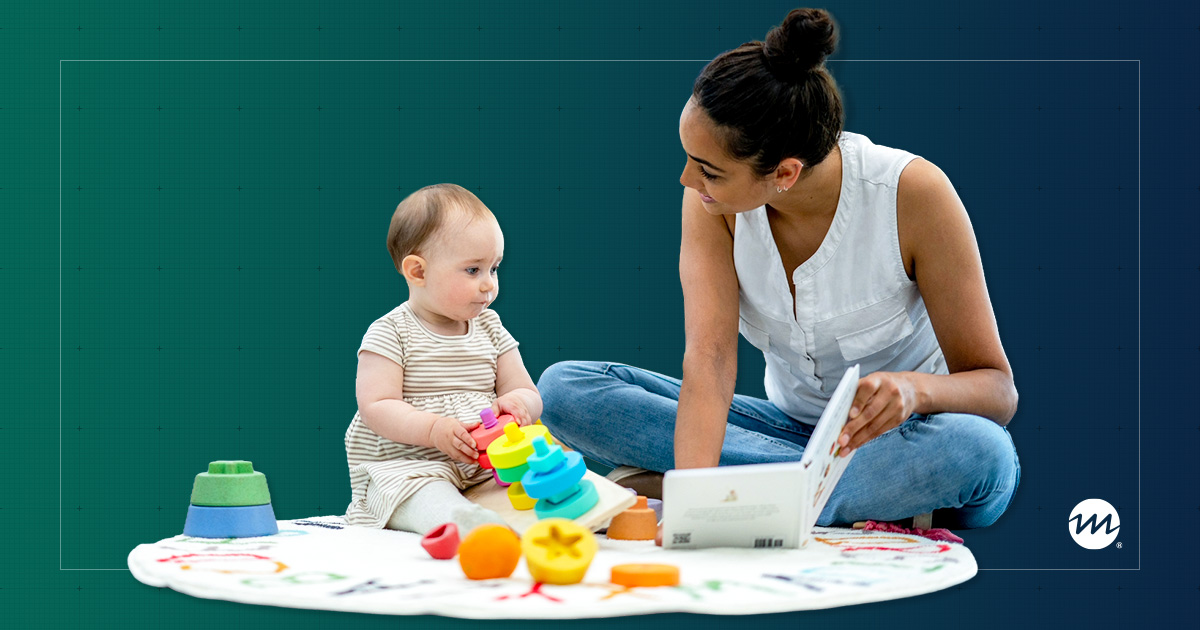 A woman sits with a baby on a playmat, surrounded by colorful toys. The baby focuses on stacking rings, while the woman holds a book. 