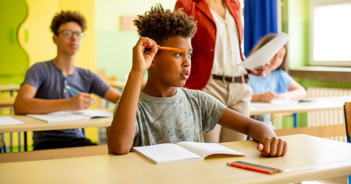 African American schoolboy is sitting in classroom and listening to lecture. In front of him are notebook and colored pens.