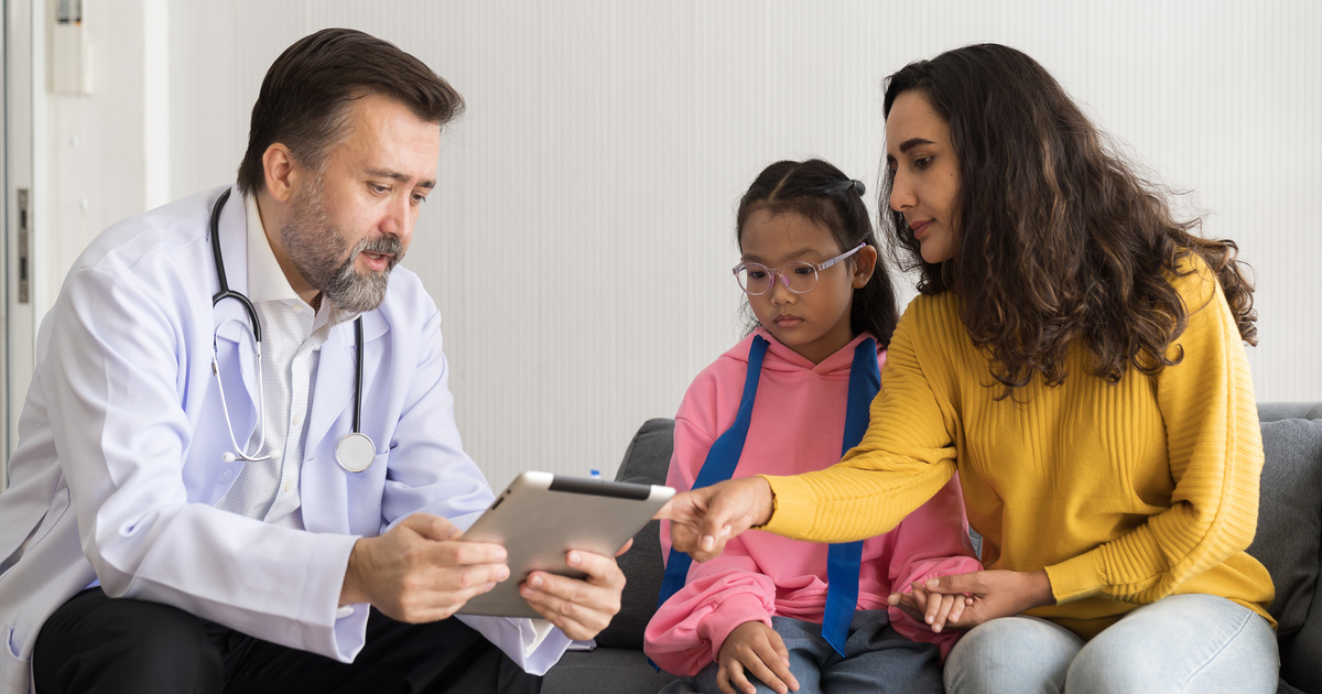 Senior male doctor and mother of child girl using digital tablet together on sofa at hospital.